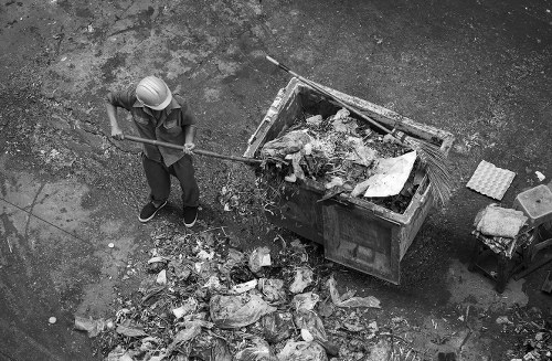 A person wearing a safety helmet and casual clothing is using a long-handled tool to clear a pile of mixed waste and rubbish from the ground next to a large industrial skip filled with debris. The debris consists of various materials such as cardboard, plastic, and packaging, with some items stacked on the pavement and others inside the skip. The scene is set outdoors on a rough, paved surface, with scattered rubbish around the skip, which is placed on a street or alleyway typical of urban areas. The image highlights the process of waste collection or clearance, likely in a setting requiring professional rubbish removal services such as those offered by Garden Clearance Paddington, serving local residents and businesses in the Paddington area. The environment appears clean and organized, with the worker focused on efficiently managing the waste, emphasizing the importance of proper rubbish disposal and clearance in urban environments.