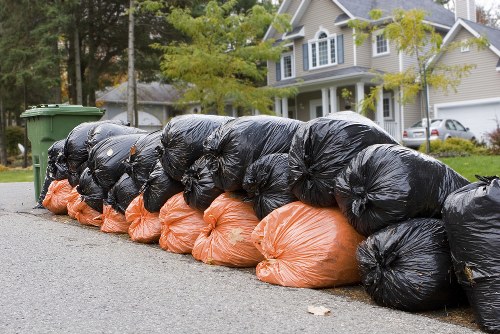 A row of tightly packed black and orange rubbish bags lined up along the edge of a residential street, with the black bags positioned behind the orange ones. The bags are filled with household waste and appear to be made of plastic, with some bags slightly bulging due to their contents. They are situated on a paved driveway or pavement area in front of a suburban property with a well-maintained lawn, trees, and a large, multi-story house with light-colored exterior walls, multiple windows, and a pitched roof. There are green wheelie bins positioned further along the street in the background. The scene suggests a scheduled rubbish collection in a typical suburban neighbourhood, with the presence of waste indicating clearance services provided by companies such as Garden Clearance Paddington, serving areas including Paddington or nearby postcode districts.