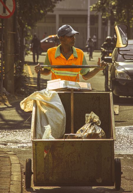 A small outdoor storage area on a paved street corner, surrounded by a chain-link fence attached to a brick wall and a building facade, contains various waste and materials typical of private rubbish