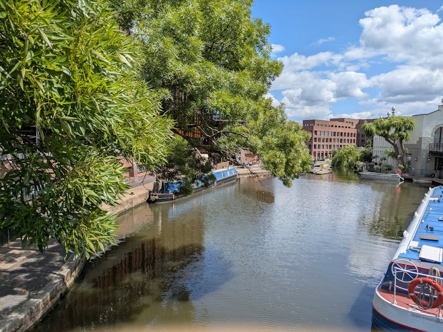 A narrowboat moored along a canal in Little Venice, featuring a prominent sign on its side that reads 'Barton Mead Little Venice.' The boat's exterior is painted in a dark colour with a red stripe alo