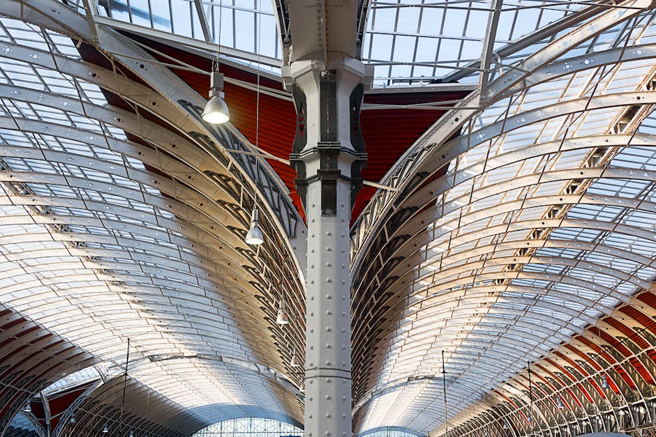 A detailed view of the arched entrance of Paddington Station featuring an ornate metal framework with decorative lattice and scrollwork. The top of the arch displays a classical crest with a shield, b