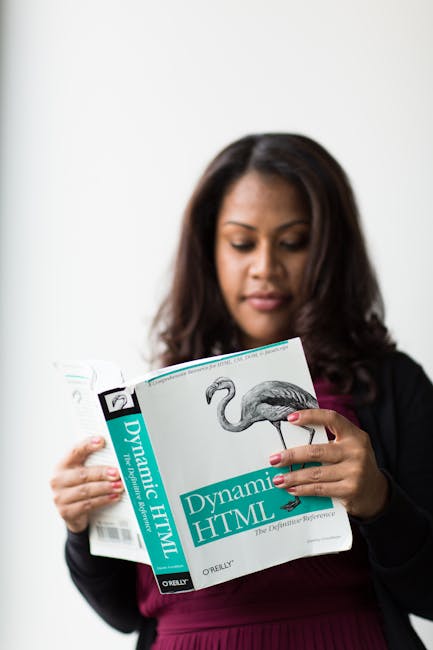 A woman with dark brown hair and medium skin tone is standing indoors against a plain white wall. She is wearing a dark blazer over a maroon top and is holding open a book titled 'Dynamic HTML' by O'R