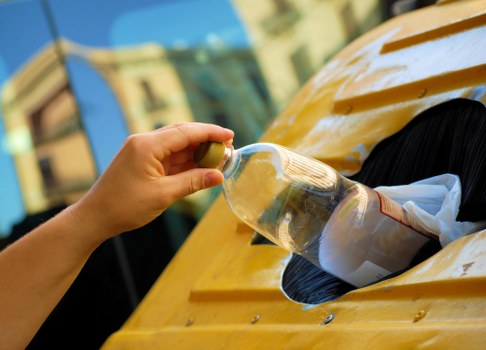 A close-up view of a person's hand holding a clear plastic bottle, filled with transparent liquid, as it is positioned above an opening in a yellow vehicle or waste container. The container appears to be made of metal with a glossy, painted yellow finish, and features a black rubber gasket around the opening. The background includes blurred architectural elements, suggesting an urban or residential setting, possibly in Paddington. The image reflects waste disposal or rubbish removal activities, with the focus on the careful handling of the bottle during rubbish collection carried out by Garden Clearance Paddington, a company specialising in rubbish and waste clearance services in the local area.