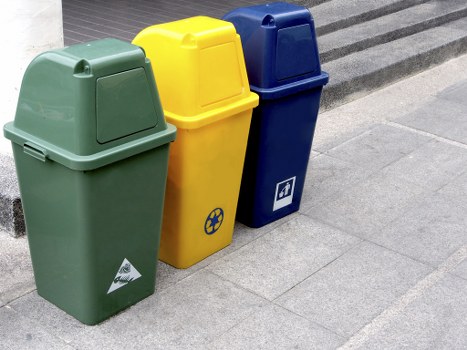 Three upright recycling bins are positioned on a paved sidewalk, adjacent to a building with dark paving stones. The first bin, on the left, is green with a slightly rounded top and a small label on the front indicating it is for waste collection, typically used for general rubbish. The middle bin is bright yellow, also with a rounded top, featuring a recycling symbol on the front, likely designated for paper or packaging waste. The third bin, on the right, is dark blue with a similar shape and a symbol on its front that suggests it is for recyclable plastics or other specific waste streams. All three bins are made from durable plastic with smooth surfaces and are situated close together, aligned in a neat row. The grey pavement beneath them appears clean, and the background includes the edge of the building or possibly a staircase, indicating an urban or town centre environment, consistent with locations in Paddington. The arrangement suggests a public waste disposal or recycling station, as managed by Garden Clearance Paddington, supporting effective rubbish removal services in the area.